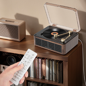 Tourne-disque de style vintage sur une surface en bois. Couvercle en plastique transparent, plateau tournant visible. Télécommande à proximité. Pile de disques vinyles à côté.