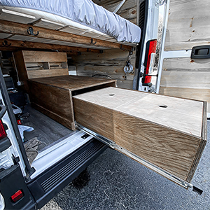 Interno di un furgone convertito, che mostra vani portaoggetti in legno e un materasso. Lo spazio è progettato per il campeggio o i viaggi.