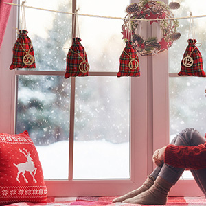 Im Fenster: rote Geschenktüten mit goldenen Verzierungen hängen an einer Schnur, darüber ein Kranz. Draußen verschneite Landschaft, drinnen ist ein roter Pullover zu sehen.