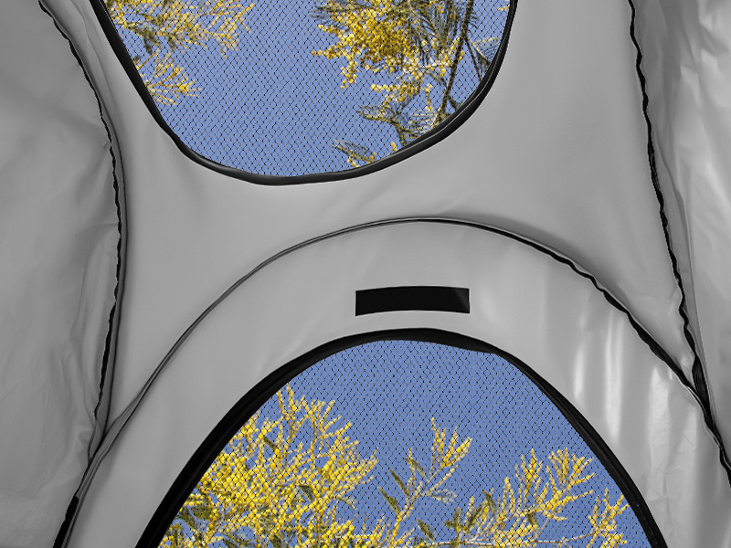 Gros plan d'une tente grise avec deux fenêtres grillagées montrant un ciel bleu et des branches d'arbres jaunes. Les fenêtres ont des sommets incurvés et des garnitures noires.