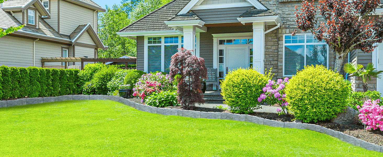 Landscaped front yard with well-maintained lawn, curved flower beds containing pink flowers and round shrubs, leading to a covered entrance with