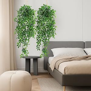 Le texte se lit comme suit : « Image 1 ». Intérieur de chambre moderne avec lit gris, pouf beige et deux grandes plantes artificielles en cascade sur une table d'appoint noire créant un accent naturel.
