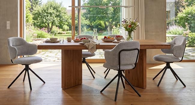 Intérieur de la salle à manger moderne avec une table ronde en bois, deux chaises rembourrées noires et grises, de grandes fenêtres donnant sur le jardin, un sol en béton et une décoration minimaliste.