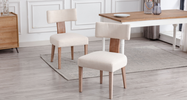 Zwei moderne Esszimmerstühle mit cremefarbener Polsterung und hellem Holzrahmen. Die Stühle verfügen über geschwungene Rückenlehnen und schmal zulaufende Beine, die auf einem grauen Teppich in der Nähe eines Holztisches positioniert