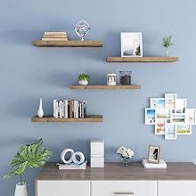 Four wooden floating shelves on blue wall. Decorated with books, plants, frames, and decor items. White cabinet below.