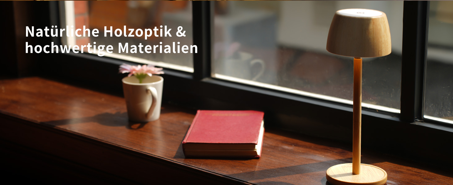 Der Text lautet „Natürliche Holzoptik & hochwertige Materialien“. Schreibtisch aus Holz mit Tischlampe und kleinem roten Buch vor einem Fenster mit Tageslicht.