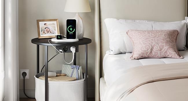 Escena de mesita de noche para dormitorio con una mesa de almacenamiento cilíndrica blanca y negra con lámpara, cargador de teléfono y objetos decorativos junto a una cama con almohadas rosas y ropa de cama blanca.