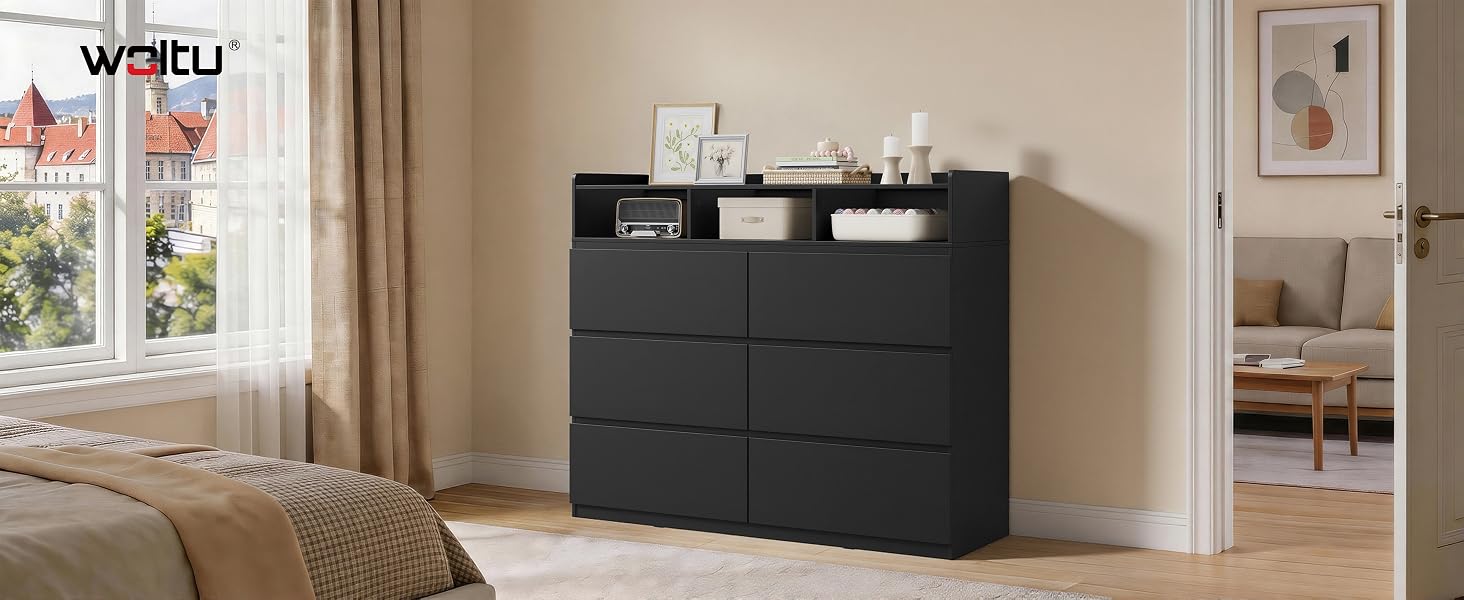 Il testo dice «WAITU». Comò nero con sei cassetti e ripiano superiore aperto, posizionato nella camera da letto beige con opere d'arte incorniciate sulla parete e scaffale bianco visibile accanto.