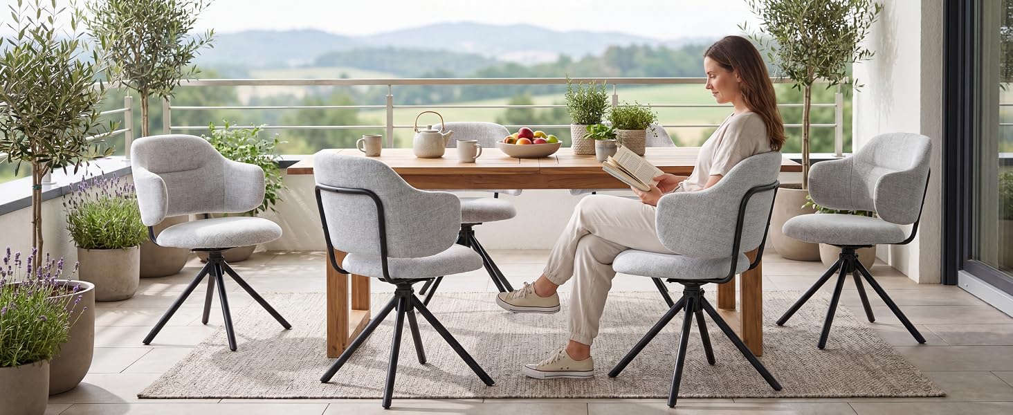 Scène de patio extérieur avec une femme assise à une table ronde avec deux chaises à pieds en métal noir, vue sur le paysage en arrière-plan, coussins décoratifs sur les meubles.
