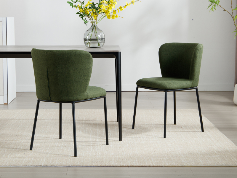 Dos sillas de comedor tapizadas en verde con respaldos curvos y patas de metal negro, colocadas cerca de una mesa oscura con un jarrón de flores amarillas