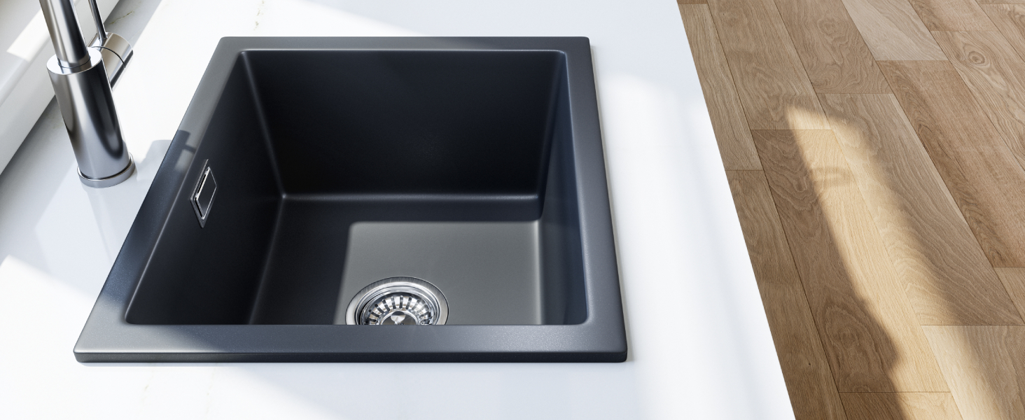Square black granite kitchen sink with single basin, set into white countertop. Stainless steel drain visible. Adjacent wooden flooring and chrome faucet partially shown.