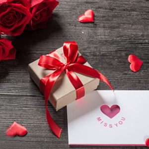 Valentine's themed composition with kraft paper gift box tied with red ribbon, red rose, scattered heart shapes, and greeting card on wooden surface.