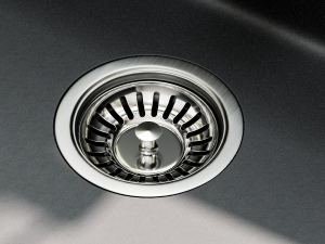 Close-up of a stainless steel kitchen sink drain. Circular design with a basket strainer insert visible. Surrounding sink surface appears to be dark-colored.