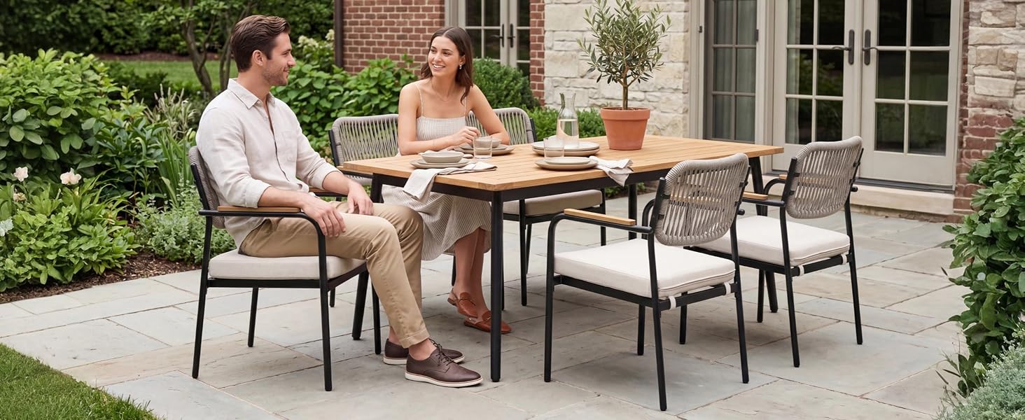 Exposition de mobilier d'extérieur : deux personnes assises à une table en bois avec des chaises modernes grises dans un jardin. Feuillage vert, plantes en pot et extérieur de la maison visibles. Configuration de salle à manger contemporaine sur la terrasse.