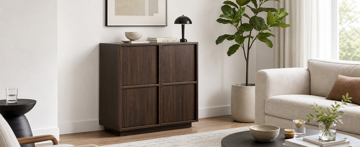 Armoire en bois foncé avec détails de panneaux géométriques positionnés dans un salon lumineux près de la fenêtre. Objets de décoration blancs et plantes en pot sur des étagères, fauteuil de couleur claire visible en arrière-plan.