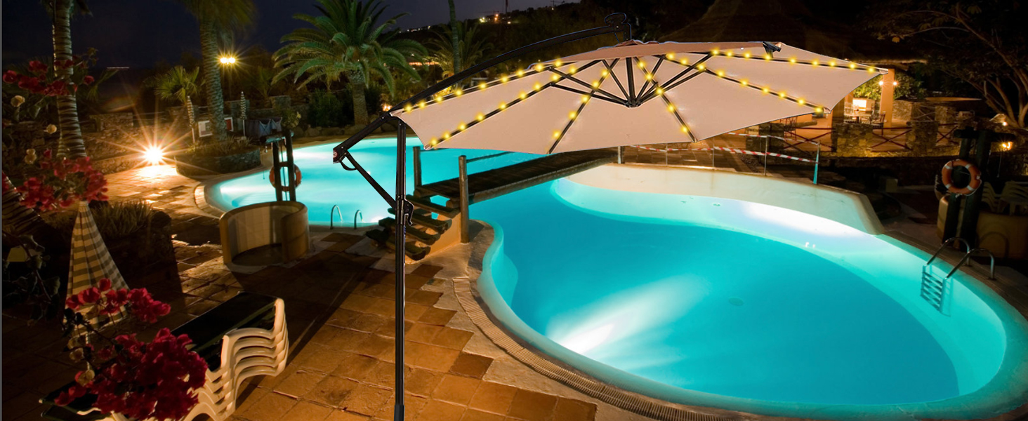 Illuminated curved swimming pool at night with turquoise water, surrounded by lounge chairs and covered by a large cream-colored umbrella.