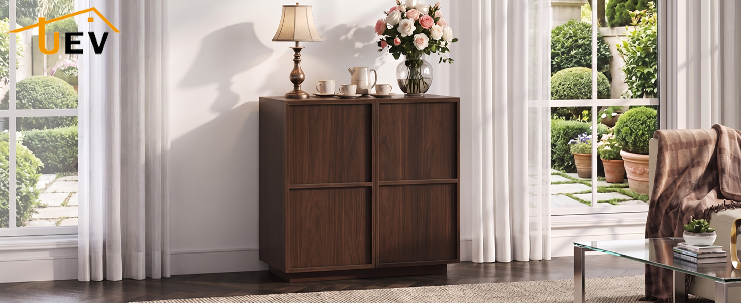 Armoire en bois foncé avec deux portes et poignées décoratives en laiton dans un salon lumineux avec de grandes fenêtres, élégante chaise rembourrée visible à côté.