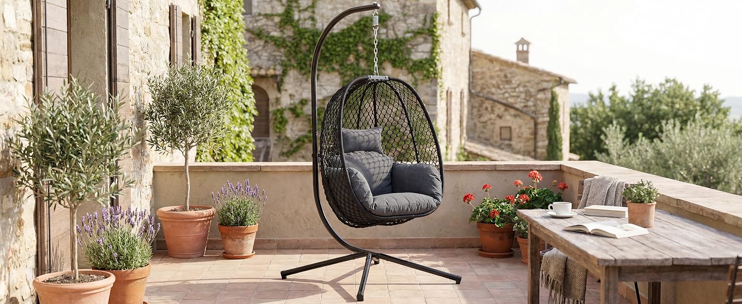 Fauteuil à œufs suspendu avec assise en forme de nacelle tissée noire et cadre en métal, positionné sur une terrasse extérieure avec des plantes en pot, des murs en pierre et des meubles de jardin visibles.