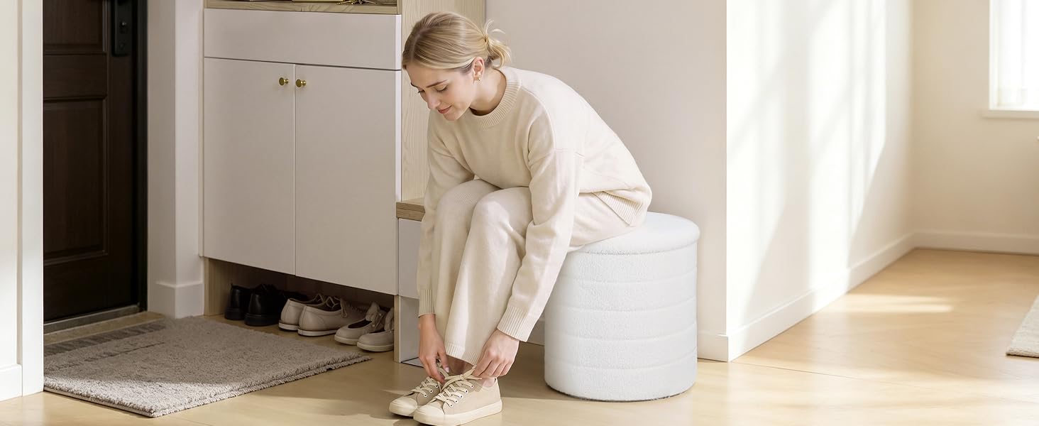 Frau in cremefarbenem Strickpullover sitzt auf einem weißen zylindrischen Hocker oder einer Ottomane im minimalistischen Innenraum mit weißen Wänden und Holzböden.