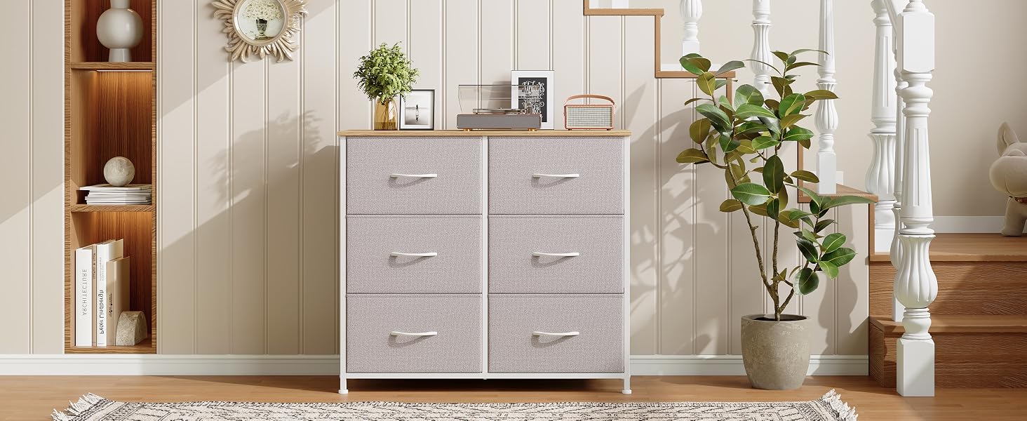 Commode en tissu gris clair avec six tiroirs et plateau en bois, posée contre un mur beige. Étagère en bois blanc visible à gauche, plante en pot à droite
