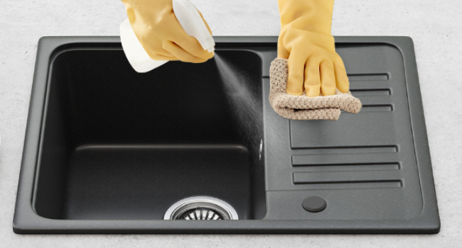Hands in yellow gloves cleaning a dark gray kitchen sink.
[SENTENCE 12] One hand sprays cleaner while the other scrubs with a sponge.
[SENTENCE 13] The sink has a single basin and a grooved draining area.