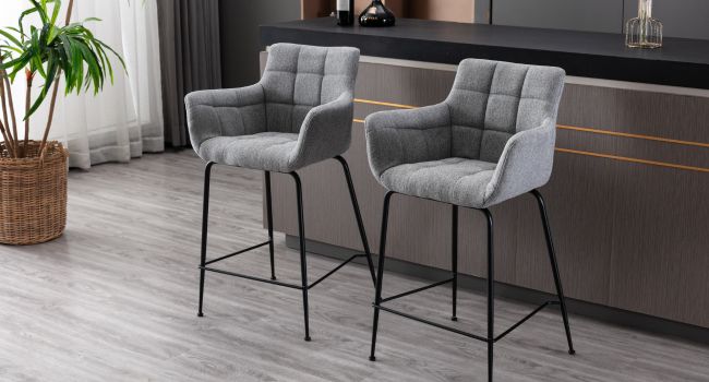 Deux tabourets de bar rembourrés gris avec accoudoirs et dossiers capitonnés. Les tabourets sont dotés de pieds en métal noir et sont placés sur un comptoir de cuisine ou un bar.
