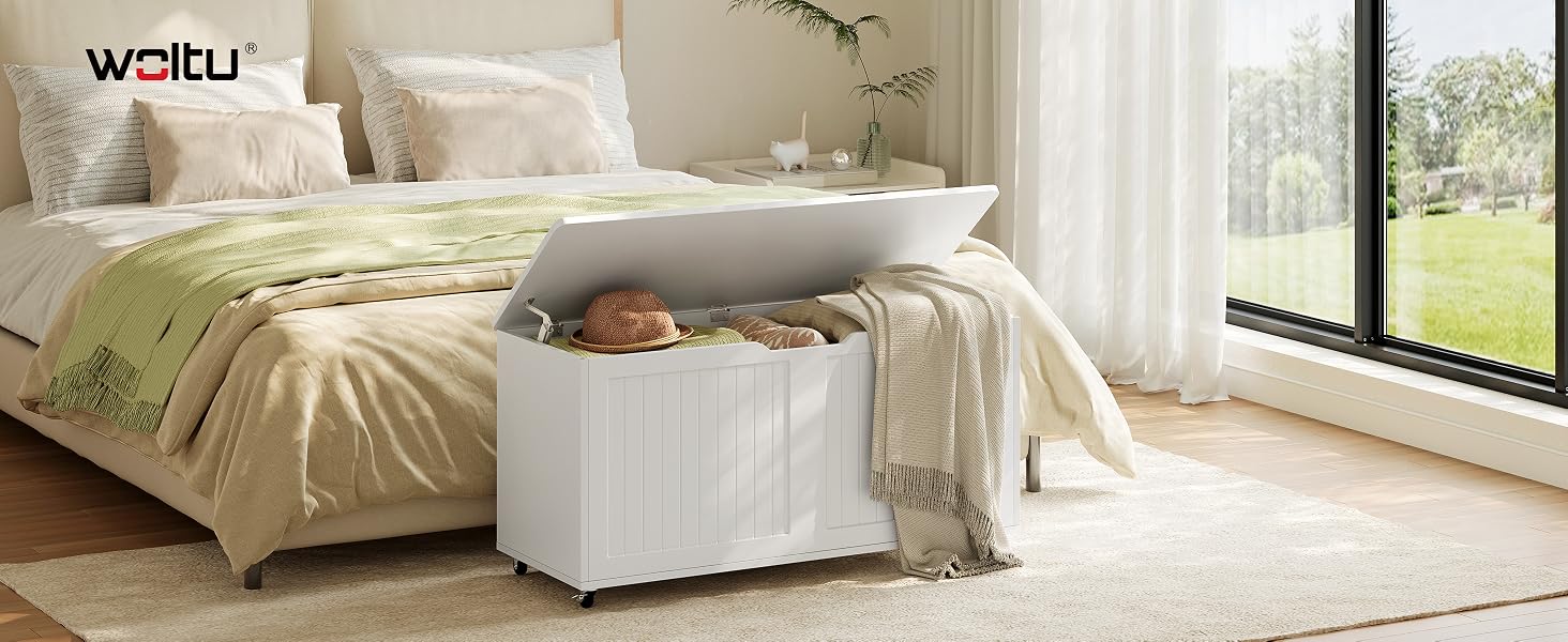 Il testo recita «WOLTU». Pouf o panca portaoggetti bianca posizionata ai piedi del letto in una luminosa camera da letto.