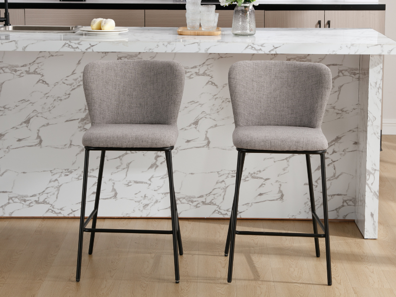 Zwei grau gepolsterte Barhocker mit geschwungener Rückenlehne und schwarzen Metallbeinen, positioniert auf einer Kücheninsel aus weißem Marmor. Minimalistisches Design, geeignet für moderne Innenräume