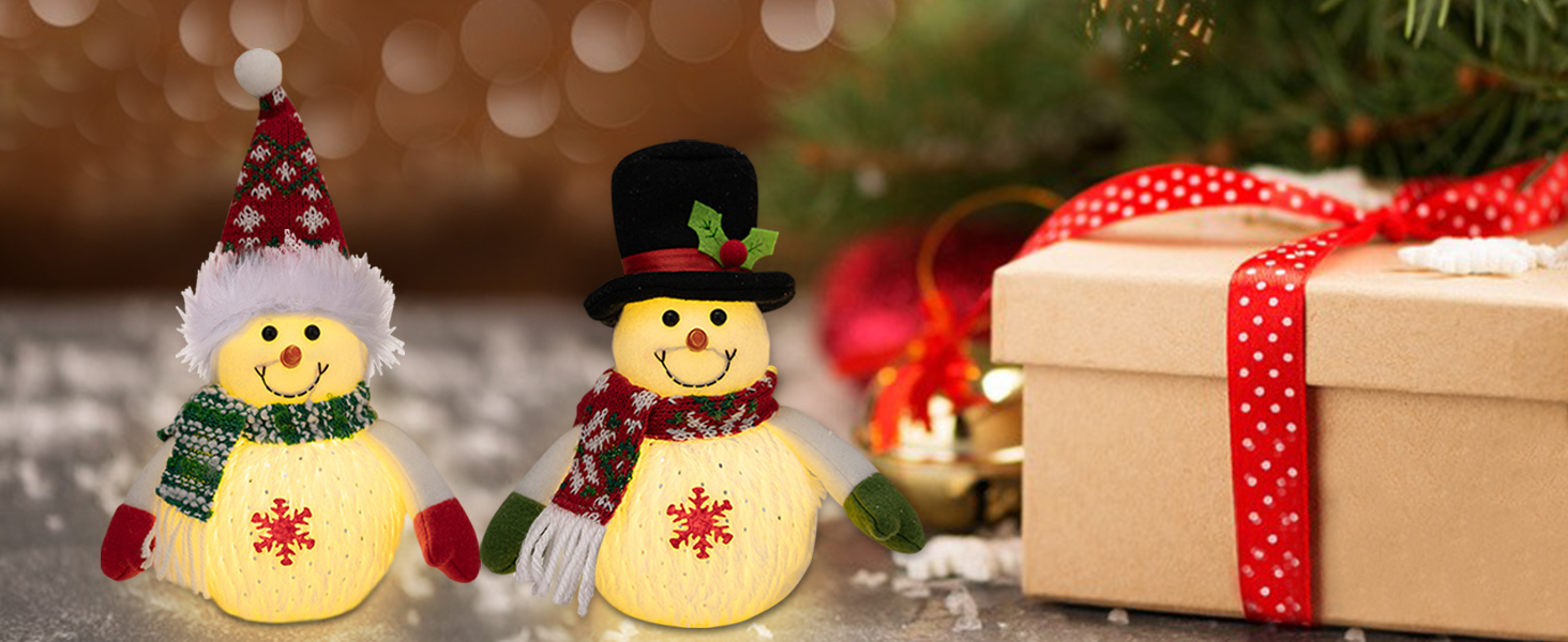 Zwei beleuchtete Schneemanndekorationen mit Strickmützen und Schals neben einer verpackten Geschenkbox mit rotem Band auf einer glitzernden Superficie