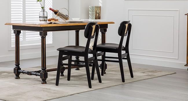 con mesa rectangular con tapa de madera y patas de metal negro. Dos sillas de madera negras con asientos acolchados. Colocadas sobre una alfombra clara contra una pared blanca con revestimiento de madera