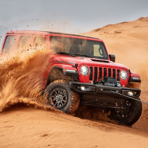 Jeep rossa che guida attraverso la sabbia del deserto, sollevando una nuvola di polvere durante un'avventura fuoristrada contro il paesaggio arancione del deserto.
