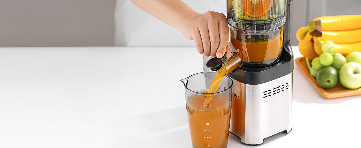 Edelstahl-Saftpresse in Betrieb, Orangensaft wird in ein Glas gegossen, im Hintergrund sind frische Früchte sichtbar