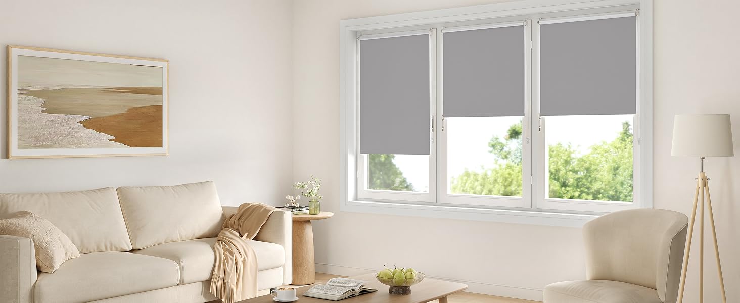 Interior moderno de sala de estar con persianas enrollables grises en ventanas triples, sofá beige, obras de arte costero e iluminación natural.