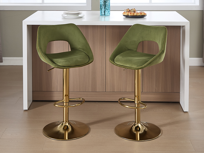 Set di moderni sgabelli da bar in velluto verde con basi cilindriche dorate, disposti in un ambiente da cucina contemporaneo con ripiani bianchi e pavimento neutro.