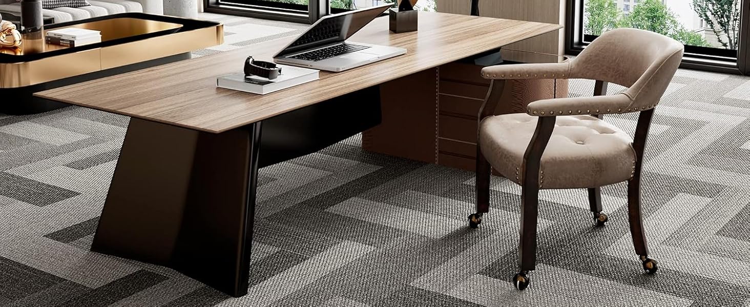 Espace de bureau moderne avec un long bureau en bois, une chaise et une installation informatique. Les grandes fenêtres fournissent un éclairage naturel. Tapis gris à motifs et plantes d'intérieur visibles.