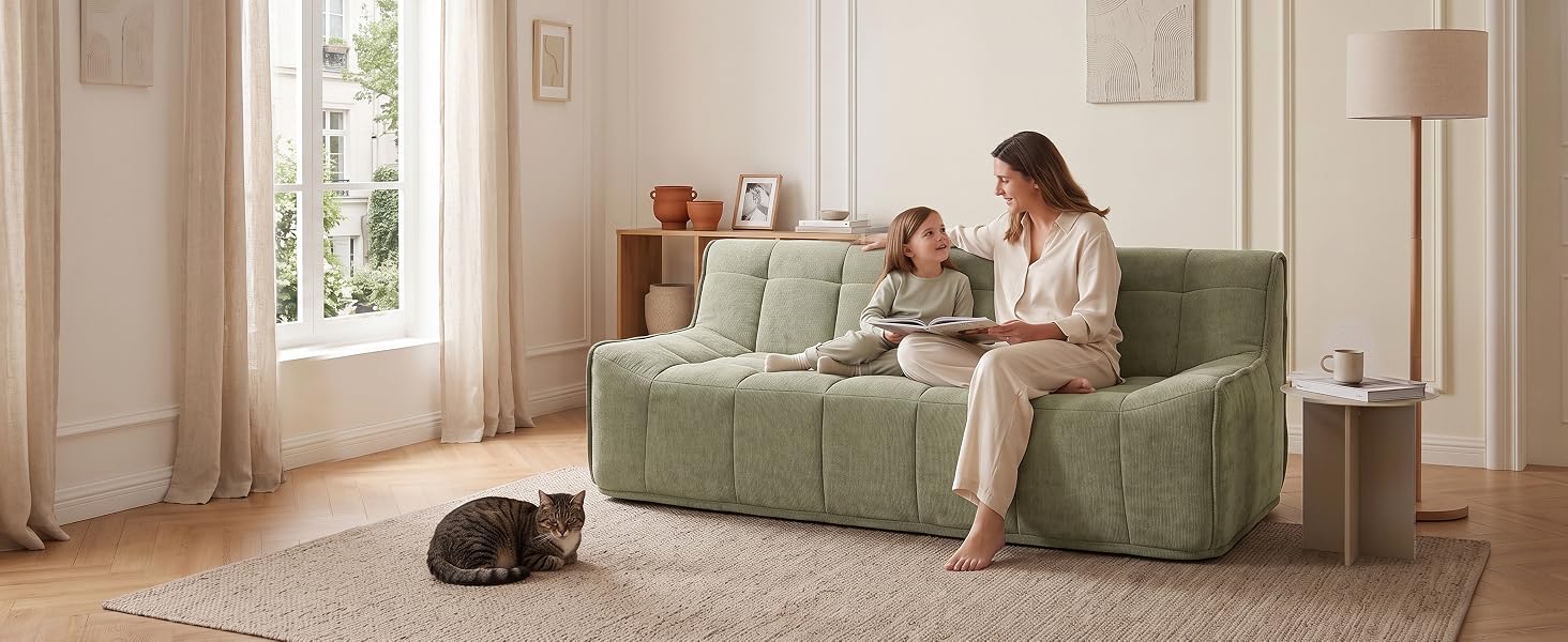 Femme assise sur un fauteuil texturé vert dans un salon lumineux et minimaliste avec des murs blancs, de grandes fenêtres et un mobilier neutre.