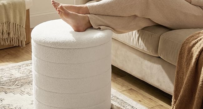 White cylindrical ottoman with ribbed texture. Person wearing beige pants resting feet on ottoman next to cream upholstered sofa with throw blanket in neutral living room setting.