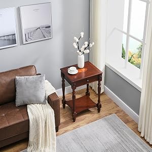 Wohnzimmerecke mit brauner Ledercouch, hölzernem Beistelltisch, weißer Vase, gerahmter Wandkunst und Fenster mit Vorhängen. Graue Wände und Holzböden sichtbar.