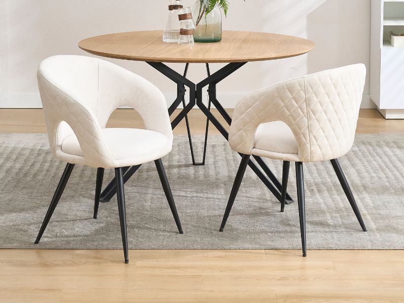 Ensemble de salle à manger moderne avec table ronde en bois avec base en forme d'étoile en métal noir, associé à deux chaises rembourrées crème matelassées sur des pieds en métal noir.