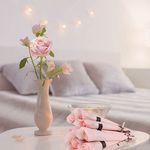 Bedroom scene with vase of pink roses, pink handheld device on bed. String lights and soft furnishings create cozy atmosphere.