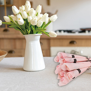 White ceramic vase filled with fresh white tulips, alongside folded pink cloth napkins on wooden surface.