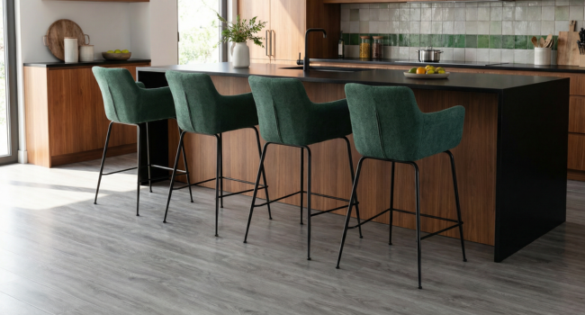 Kitchen island with emerald green velvet bar stools, dark countertop, and wooden base cabinetry. Grey flooring and glass backsplash visible.