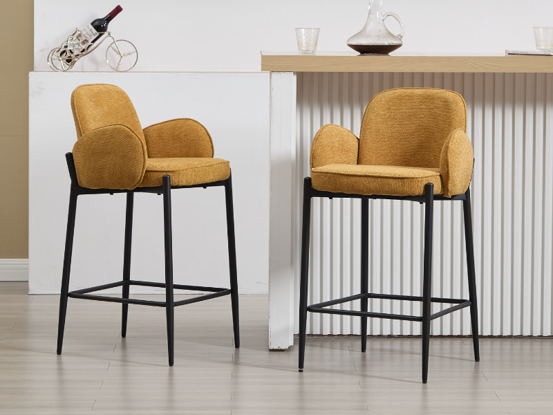 Dos taburetes de bar modernos con asientos y respaldos tapizados en amarillo mostaza, con marcos y reposapiés de metal negro, comparados con un ambiente interior neutro con mostrador.