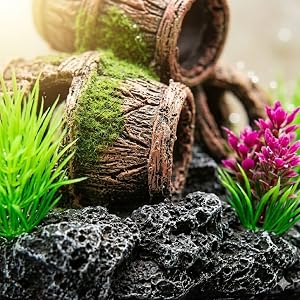 Aquarium decoration scene with wooden barrel, vibrant green artificial plants, pink flowering plant, and black rocky substrate.