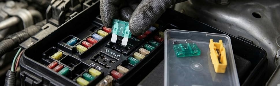 Gloved hand installing or working with a small electronic component on a dark circuit board or automotive component with yellow battery connector visible.