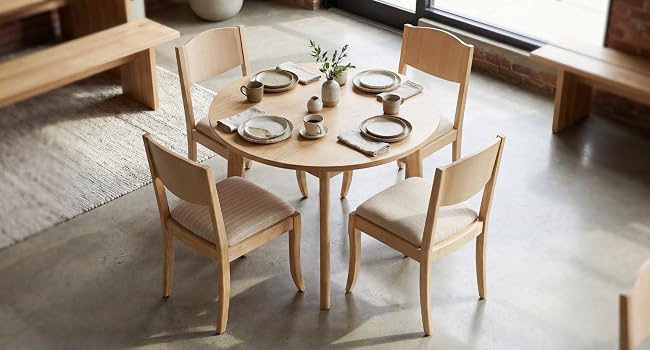 Table à manger en bois clair avec deux chaises assorties dans un espace intérieur minimaliste. Petite figurine en robe rouge positionnée sur la surface de la table.