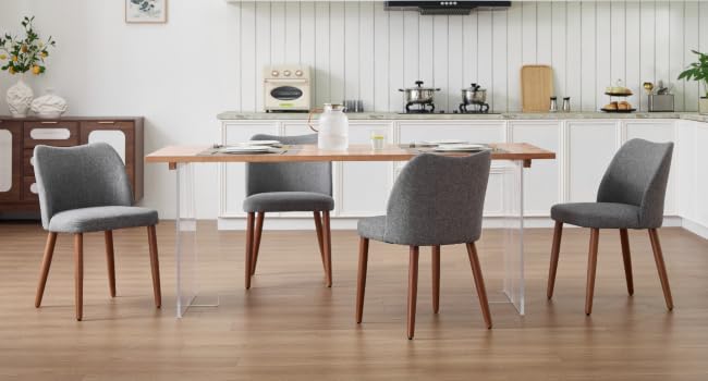 Modern dining area with four gray upholstered chairs around wooden table, white walls, and minimal decor with plants in background.