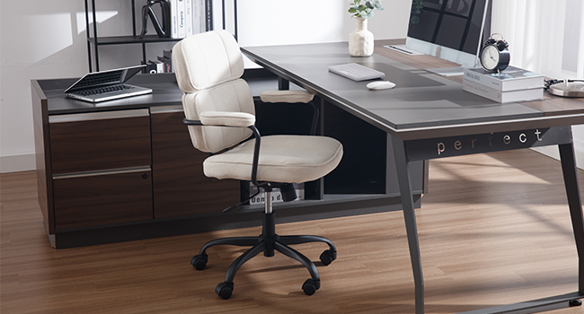 L-shaped desk with dark wood finish and gray top. White ergonomic office chair. Desktop computer, books, and plant visible. Modern home office setup on hardwood floor.