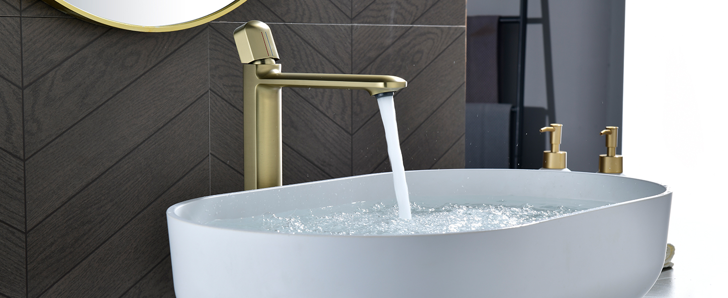avec finition dorée brossée, versant de l'eau dans un lavabo ovale blanc. Fond de mur carrelé foncé avec des luminaires dorés supplémentaires visibles.