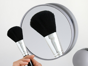 Two makeup brushes with white handles and black bristles, shown next to a round mirror. One brush is being held by a hand.